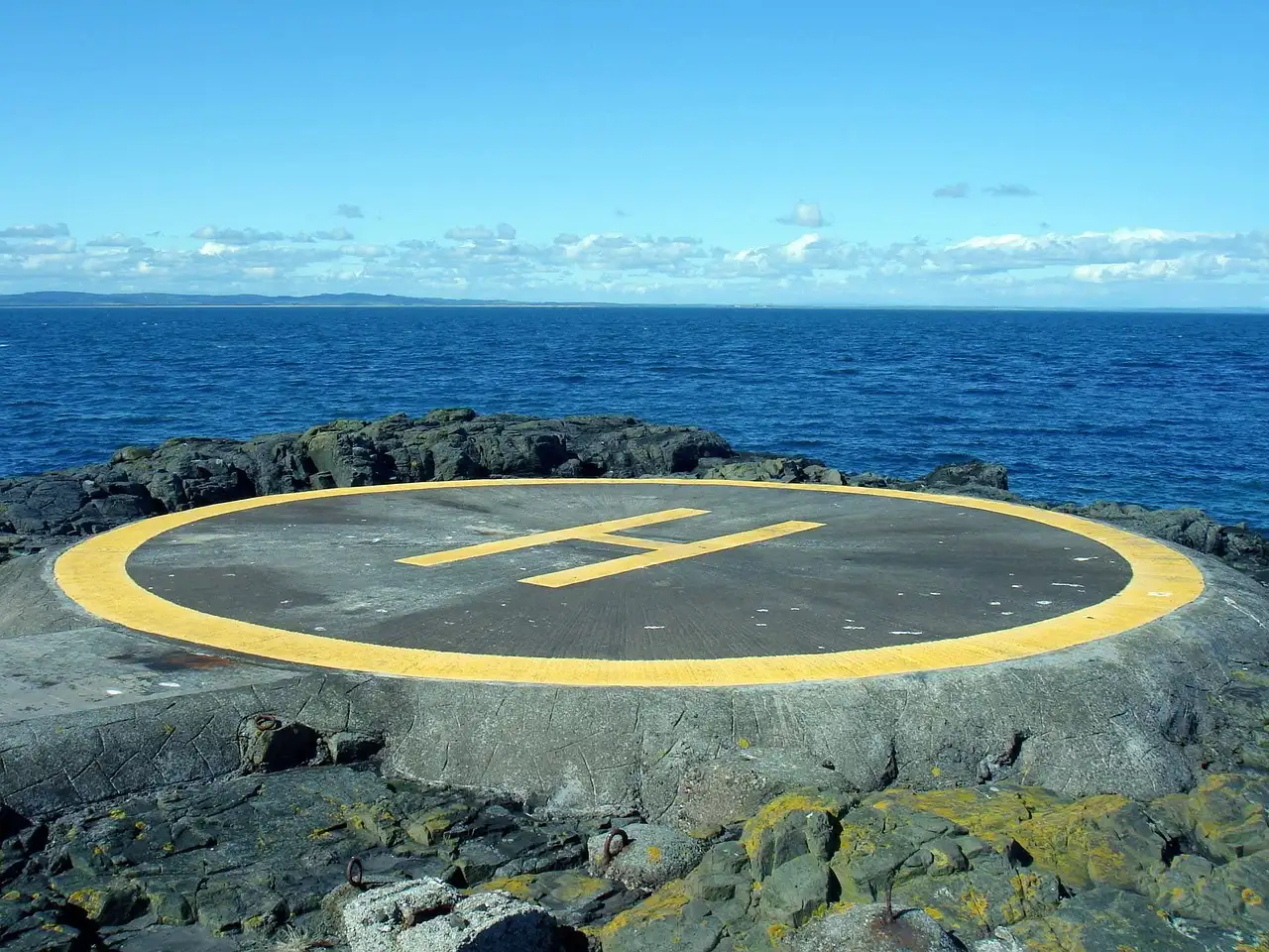 A round yellow helicopter landing pad on dark rocks by a blue sea