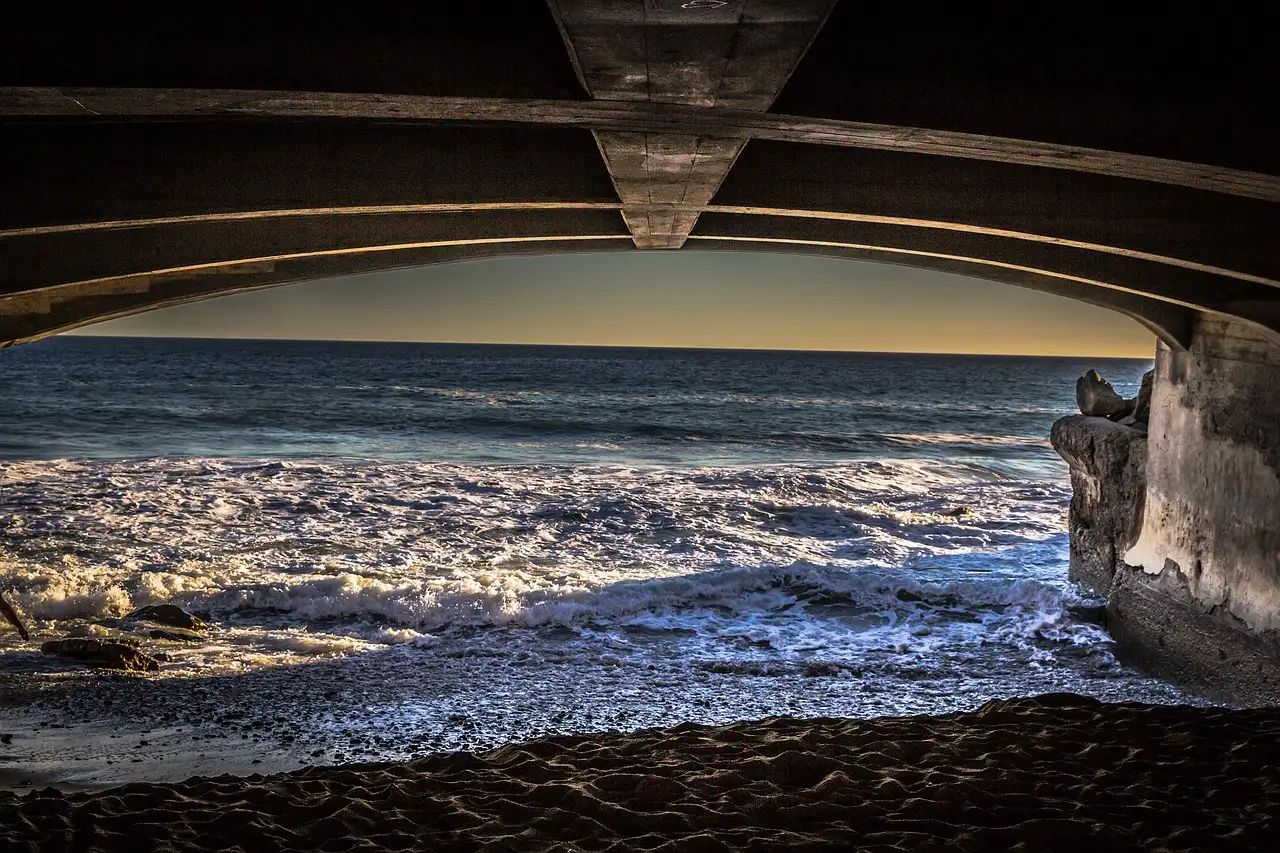 Waves rolling onto a Malibu beach, seen from under a bridge at golden hour
