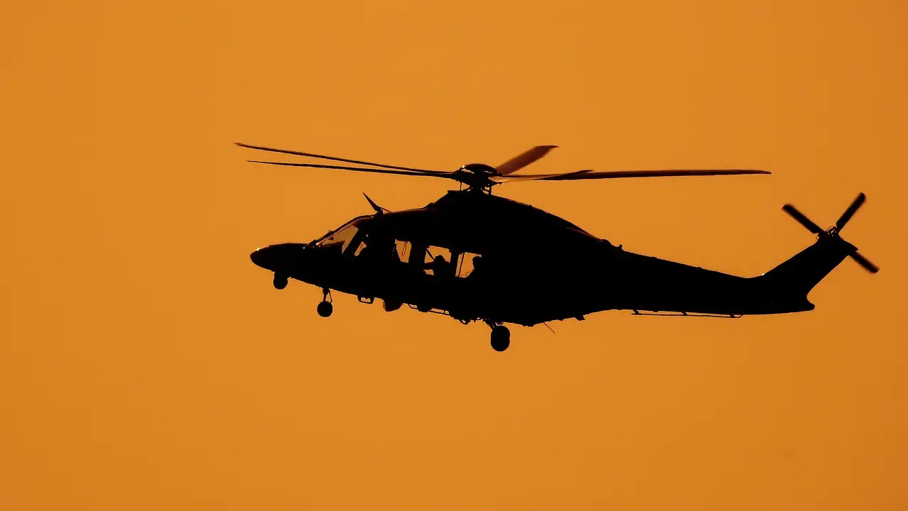 The dark outline of a helicopter flying against an orange sunset sky