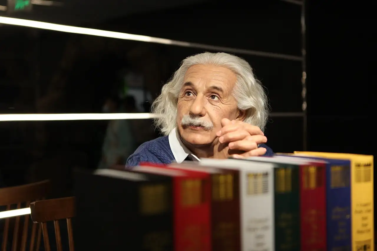 A wax figure of Albert Einstein resting his chin on his hands beside a row of books