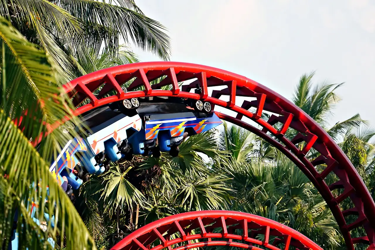 A red roller coaster track between palm trees