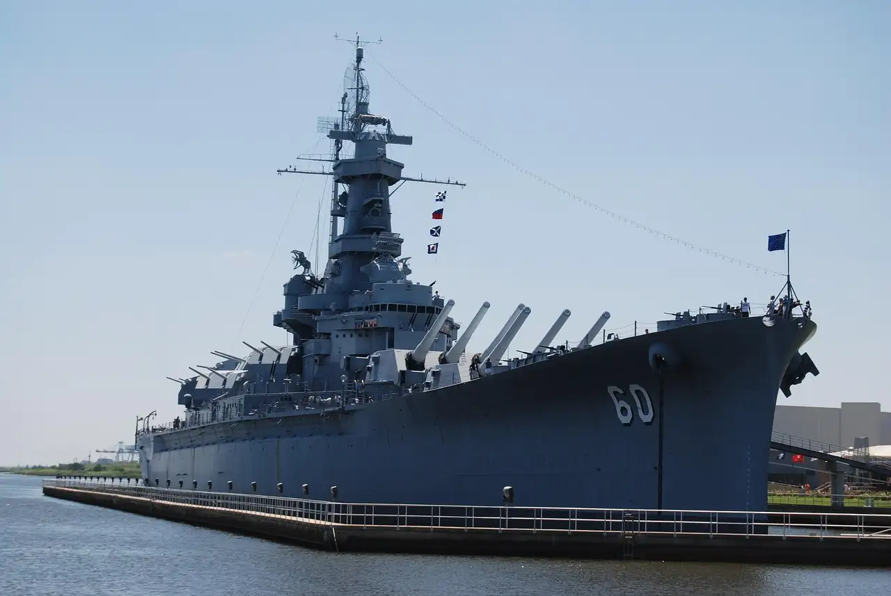 A grey battleship with big gun turrets and a tall mast, moored at a pier under a clear sky