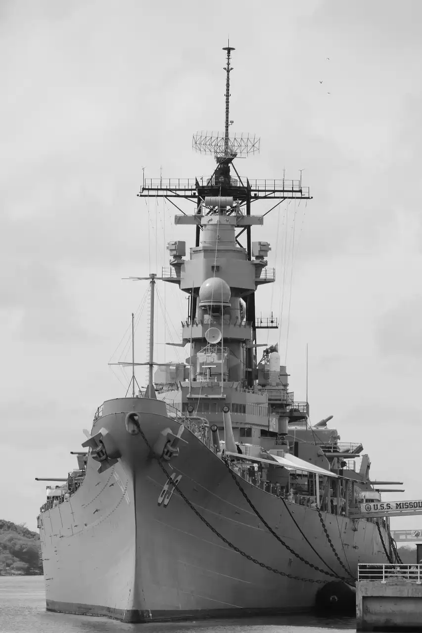 A grey Iowa-class battleship moored at a pier, seen from the bow, in black and white