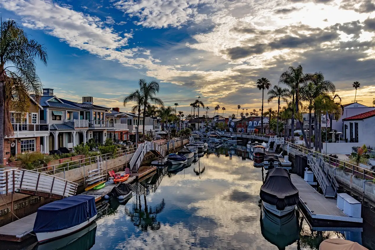 A canal in Long Beach lined with palm trees, waterfront houses and small boats, with clouds reflected in the still water