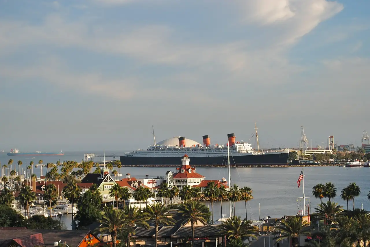 The Queen Mary ocean liner with its three red funnels, docked across the harbor from palm trees and low waterfront buildings in Long Beach