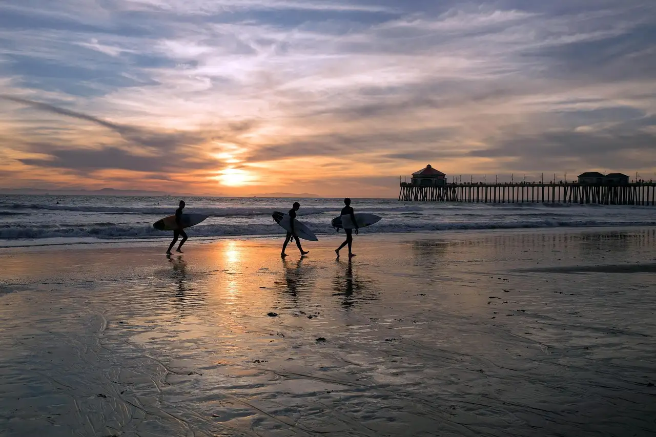 Surfers carrying boards along the beach at sunset near a pier