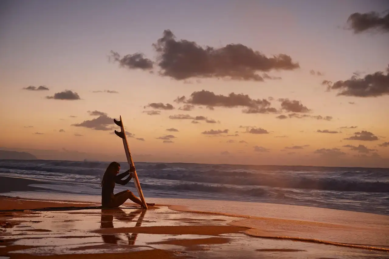 A surfer kneeling with a board on the beach at sunset